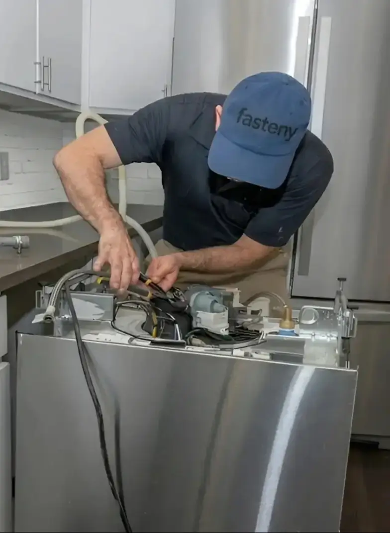 A technician from Fasteny Appliance Repair, performing expert dishwasher repair on a built-in unit in a Greater Boston Area home kitchen. The technician is focused and uses pliers on exposed components, with "fasteny" visible on his cap.