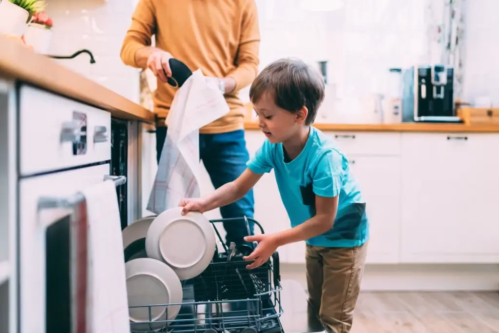 A happy son helps load clean plates into a freshly fixed dishwasher in a bright kitchen.